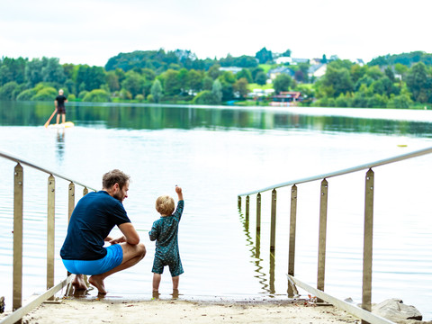 barrierefreier Badestelle an der Brucher Talsperre Ein Vater und sein Kind beobachten am Seeufer ein ruhiges Wasserpanorama mit grünen Bäumen im Hintergrund.
