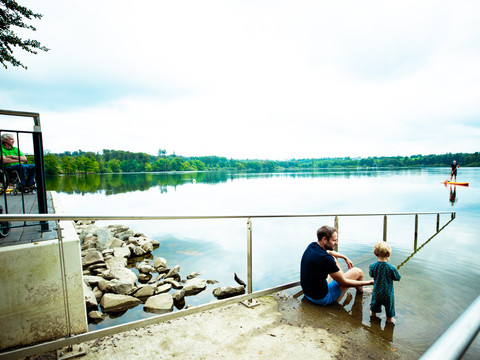barrierefreie Badestelle an der Brucher Talsperre  <p>Vater und Kind am Seeufer neben einer Rampe; im Wasser ein Stand-up-Paddler.</p>