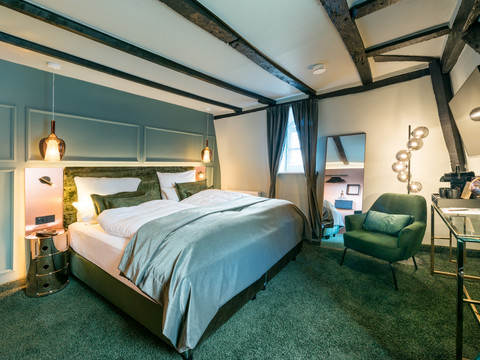 Hotel von Landsberg Gemütliches Hotelzimmer mit Holzbalken, Doppelbett, grünem Sessel, Schreibtisch und dekorativer Beleuchtung.
