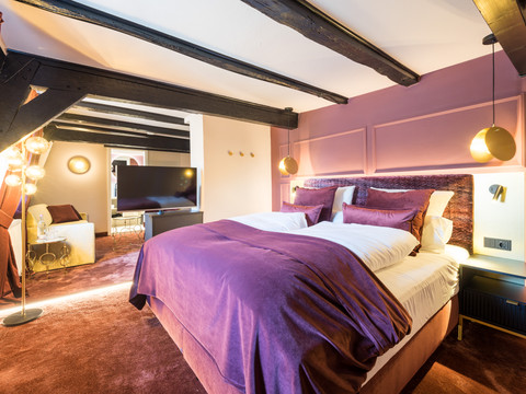 Hotel von Landsberg Elegantes Hotelzimmer mit violettem Bettbezug, modernen Lichtakzenten und sichtbaren Holzbalken.