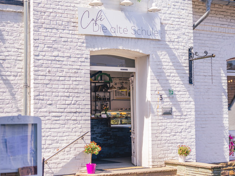 Café Die alte Schule Das Bild zeigt den Eingang des Cafés "Die alte Schule" in einem weißen Backsteingebäude mit blühendem Blumenkasten.