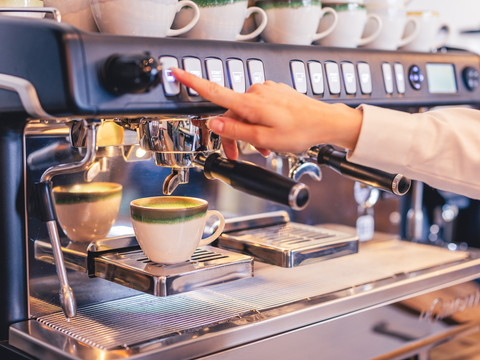 Café Die alte Schule Eine Hand betätigt eine Taste an einer modernen Espressomaschine, während eine Tasse darunter steht.