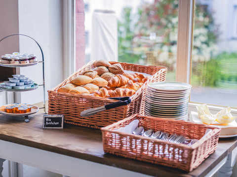 Café Die alte Schule Körbe mit frischen Brötchen und Croissants auf einem Tisch, daneben Teller und Besteck bereitgestellt.