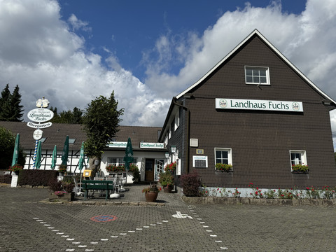 Landhaus Fuchs - Außenansicht  Das Landhaus Fuchs in Kürten mit Gastgarten und Parkplätzen vor grauem, zweistöckigem Gebäude.