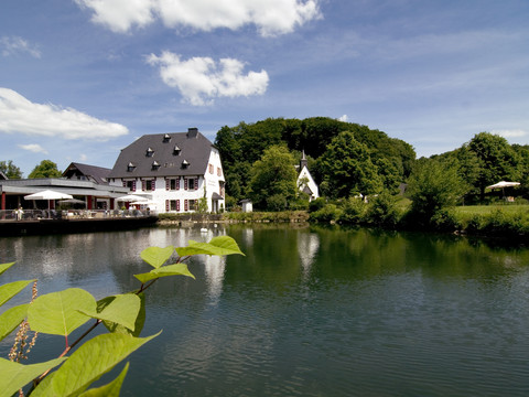 Malteser Komturei-6 Ein weißes, historisches Gebäude am See mit Terrasse; umgeben von grüner Natur und blauem Himmel.