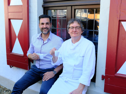 Porträt Zwei Männer sitzen vor einem Fachwerkfenster, genießen gemeinsam eine Kaffeepause im Freien.