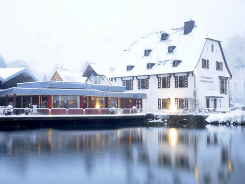 Malteser Komturei-Winter Hotel Gronau am winterlichen Seeufer in Bergisch Gladbach, mit verschneiten Dächern und warmem Licht.