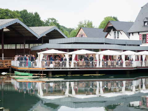 Malteser Komturei- 3 Eine Hochzeitsgesellschaft feiert fröhlich auf einer überdachten Holzterrasse über einem Teich.