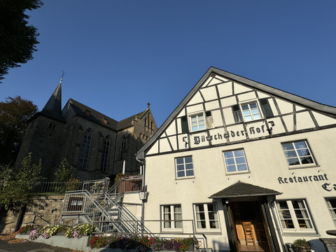 Dürscheider Hof - Außenansicht Restaurant & Kirche  <p>Fachwerkhaus "Büscheder Hof" neben einer Kirche in Kürten, unter blauem Himmel, umgeben von Blumen.</p>