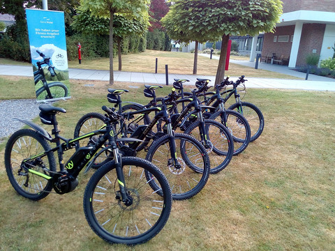 Fahrräder Sieben schwarze Fahrräder mit grünen Akzenten stehen auf einer Rasenfläche neben einer Infotafel.