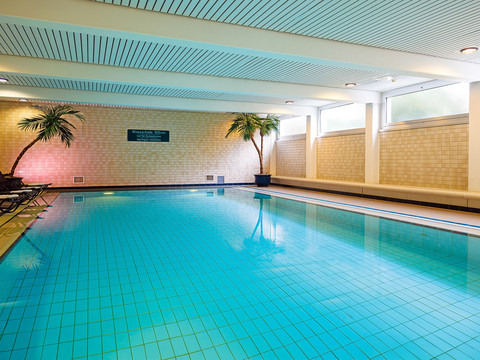 Schwimmbad Innenbereich eines Hallenbades mit türkisblauem Wasser, Liegen und zwei Palmen an den Seiten.