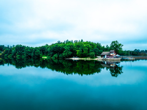 Brucher Talsperre aus der Luft Eine idyllische Seelandschaft mit Bäumen und einem Bootshaus, das sich im ruhigen Wasser spiegelt.