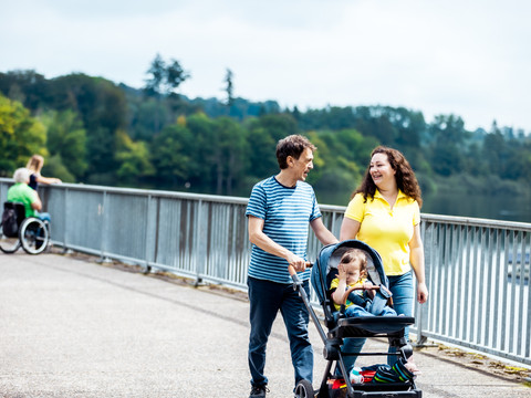 Staumauer an der Brucher Talsperre Ein Paar spaziert mit einem Kleinkind im Buggy an einem Seeufer entlang, im Hintergrund Wälder.