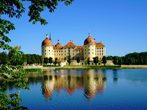 Schloss Moritzburg.jpg Schloss Moritzburg spiegelt sich in einem ruhigen See, umgeben von üppigem Grün und blauem Himmel.Moritzburg Castle is reflected in a tranquil lake, surrounded by lush greenery and blue skies.Zámek Moritzburg se odráží v klidném jezeře obklopeném bujnou zelení a modrou oblohou.Zamek Moritzburg odbija się w spokojnym jeziorze, otoczonym bujną zielenią i błękitnym niebem.Kasteel Moritzburg wordt weerspiegeld in een kalm meer, omringd door weelderig groen en blauwe luchten.Il castello di Moritzburg si specchia in un lago tranquillo, circondato da una vegetazione lussureggiante e da un cielo azzurro.