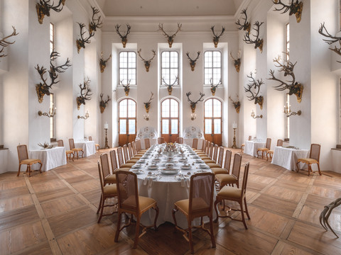 Geweihsammlung im Speisesaal.jpg Ein Speisesaal mit Kandelabern und Geweihen an weißen Wänden und einem gedeckten Tisch.A dining room with candelabras and antlers on white walls and a laid table.Jídelna se svícny a parohy na bílých stěnách a prostřeným stolem.Jadalnia z kandelabrami i porożami na białych ścianach i nakrytym stołem.Een eetkamer met kandelaars en geweien op witte muren en een gedekte tafel.Una sala da pranzo con candelabri e corna su pareti bianche e una tavola imbandita.