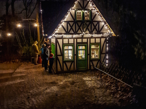 Märchenwald Winterzauber Fachwerkhäuschen nachts beleuchtet, umgeben von Bäumen, mit Menschen in Winterkleidung davor.