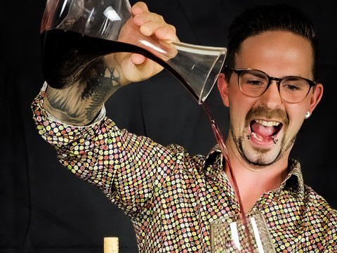 Bergischer Weinkeller Mann mit gemusterter Hemd und Brille schenkt Rotwein aus einer Karaffe in ein Glas ein.