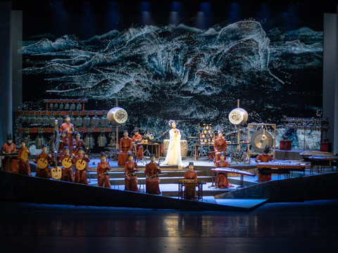 Ein Orchester in traditionellen roten Gewändern spielt auf einer Bühne vor einem dramatischen Hintergrund.An orchestra in traditional red robes plays on a stage against a dramatic backdrop.Na pódiu hraje orchestr v tradičních červených hábitech na dramatickém pozadí.Orkiestra w tradycyjnych czerwonych szatach gra na scenie na dramatycznym tle.Een orkest in traditionele rode gewaden speelt op een podium tegen een dramatische achtergrond.Un'orchestra in abiti tradizionali rossi suona su un palco con uno sfondo drammatico.