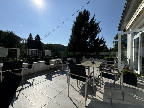 Kaffeerösterei Pagnia - Ansicht Außenterrasse Sonnige Terrasse mit Tischen und Stühlen, umgeben von Bäumen, unter klarem, blauem Himmel.