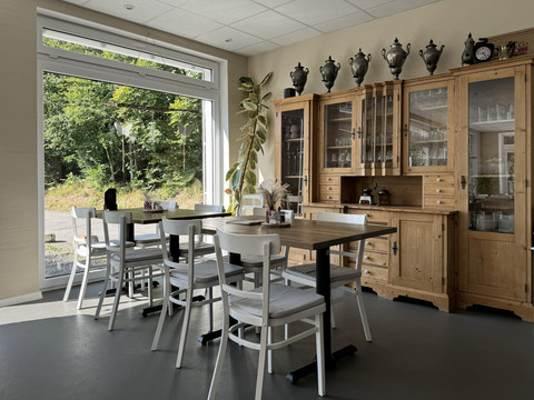 Kaffeerösterei Pagnia - Innenansicht Gemütlicher Raum mit Holzmöbeln und weißen Stühlen, großen Fenstern und einer Pflanze im Eck.