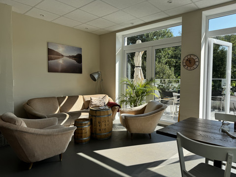 Kaffeerösterei Pagnia - Innenansicht Gemütlicher Raum mit beigen Sesseln, Sofas und Holzfässern, Blick auf sonnige Terrasse durch Glastüren.