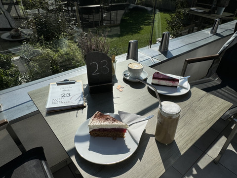 Kaffeerösterei Pagnia - Ansicht Kaffee & Kuchen Zwei Teller mit Kuchen und Getränken auf einem Tisch, Blick auf eine grüne Wiese im Hintergrund.