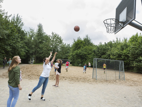 Jugendherberge Lindlar Jugendliche spielen Basketball in einem Park mit sandigem Spielfeld, umgeben von grünen Bäumen.