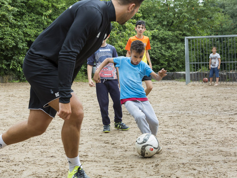 Jugendherberge Lindlar Kinder spielen Fußball auf einem sandigen Platz, umgeben von Bäumen, während ein Erwachsener zuschaut.