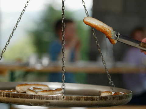 Jugendherberge Wiehl Person grillt Würste im Freien auf einer hängenden Pfanne. Hintergrund zeigt unscharfe Natur.