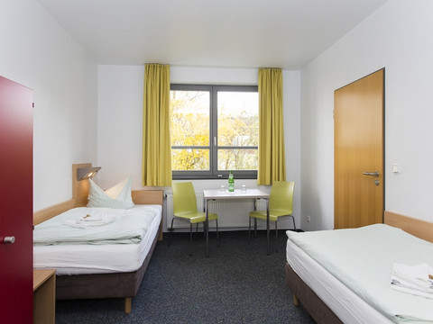 Jugendherberge Wiehl Ein helles Hotelzimmer mit zwei Betten, Tisch, zwei Stühlen, großem Fenster und gelben Vorhängen.