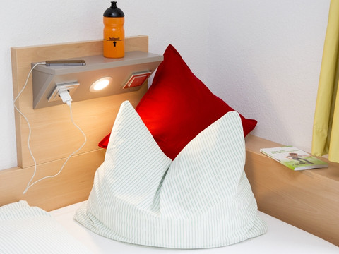 Jugendherberge Wiehl Ein gemütliches Bett mit einer leuchtend roten Kissenhülle und einer Lampe auf einem Holzbrett.