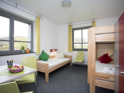 Jugendherberge Wiehl Helles Hostelzimmer mit Einzelbett, Etagenbett, Tisch und Stühlen, fröhliche Farbgebung.