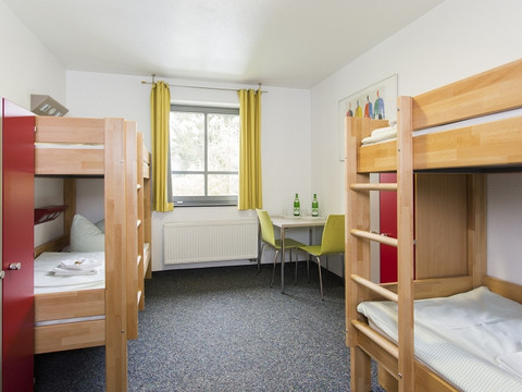 Jugendherberge Wiehl Helles Hostelzimmer mit drei Etagenbetten, Tisch und Stühlen, Fenster mit gelben Vorhängen.