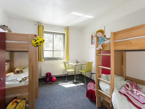 Jugendherberge Wiehl Helles Kinderzimmer mit gelben Vorhängen, Stockbetten und Spielzeug, darunter Helme und Puppen.
