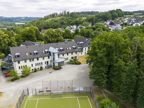 Jugendherberge Wiehl Luftaufnahme eines modernen zweistöckigen Gebäudes, umgeben von Bäumen, mit einem Sportplatz im Vordergrund.