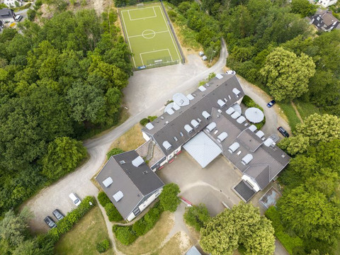 Jugendherberge Wiehl Luftaufnahme eines großen Gebäudekomplexes mit Fußballplatz, umgeben von Bäumen und Parkplätzen.