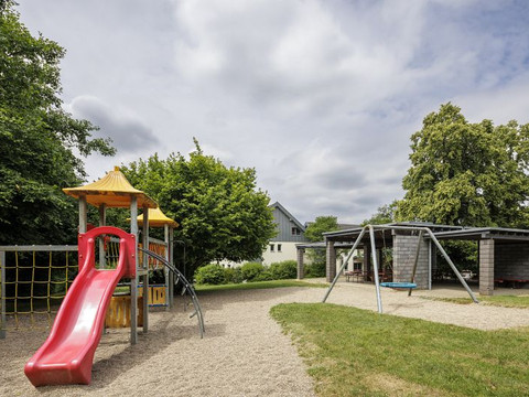 Jugendherberge Wiehl Spielplatz mit bunter Rutsche, Kletternetz und Schaukel unter Bäumen, bedeckter Himmel.
