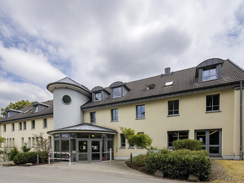 Jugendherberge Wiehl Barrierefreies Gebäude mit gelber Fassade, modernem Eingang und mehreren Dachfenstern unter wolkigem Himmel.