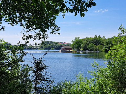Brucher Talsperre Waldsee mit Bootshaus und Wald im Hintergrund, blauer Himmel, grüne Ufervegetation, idyllische Ruhe.