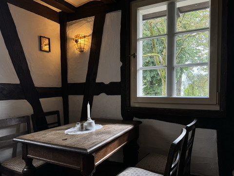 Pfannkuchenhaus Nümbrecht - Innenansicht Sitzecke Gemütliche, rustikale Ecke eines Fachwerkhauses, mit Holztisch und Bauernstühlen, Fenster zeigt Gartenblick.