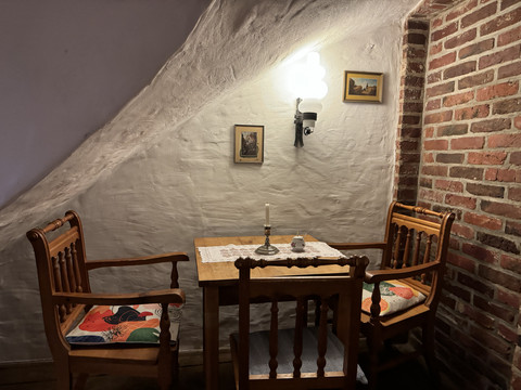 Pfannkuchenhaus Nümbrecht - Ansicht Sitznische Gemütliche Sitzecke aus Holz mit zwei Stühlen, Tisch, Kerze, unter schräger, weißer Wandbeleuchtung.