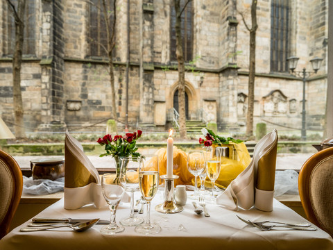 Restaurant in the Romantik Hotel Deutsches Haus Elegant gedeckter Tisch mit brennender Kerze, Sektgläsern und roten Rosen vor einem großen Fenster mit Blick auf eine alte Steinmauer.Elegantly laid table with lighted candles, champagne glasses and red roses in front of a large window with a view of an old stone wall.Elegantně prostřený stůl s hořícími svíčkami, skleničkami na šampaňské a červenými růžemi před velkým oknem s výhledem na starou kamennou zeď.Elegancko nakryty stół z płonącymi świecami, kieliszkami szampana i czerwonymi różami przed dużym oknem z widokiem na stary kamienny mur.Elegant gedekte tafel met brandende kaarsen, champagneglazen en rode rozen voor een groot raam met uitzicht op een oude stenen muur.Tavola elegantemente apparecchiata con candele accese, calici di champagne e rose rosse davanti a una grande finestra con vista su un antico muro di pietra.