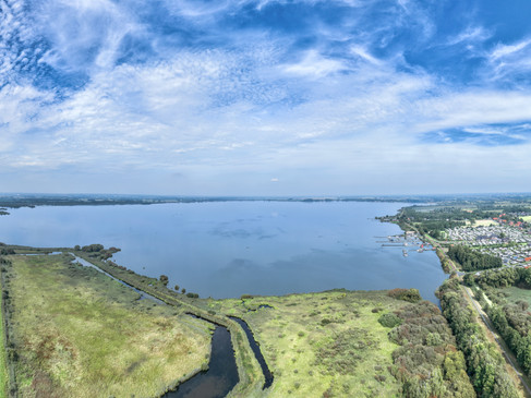 Weitläufige Seelandschaft mit umliegenden Wiesen und Wäldern unter einem blauen Himmel.