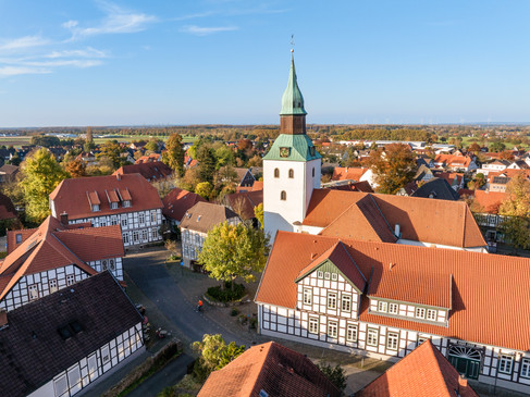 Kirchturm mit grünem Dach überragt Fachwerkhäuser in herbstlicher Dorfansicht.