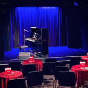Theater509 / Bürgerhaus Stollwerck Ein gemütlicher Theatersaal mit roten Tischdecken, Stühlen, einem Piano und einem stehenden Mikrofon.A cozy theater hall with red tablecloths, chairs, a piano and a standing microphone.