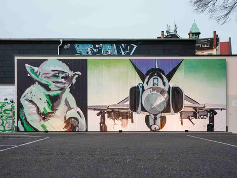 04_KSC_Collateral Sculptures_Doll_Das Licht.jpg Ein Wandgemälde zeigt Yoda links und ein Kampfflugzeug rechts vor einem Gebäude.A mural shows Yoda on the left and a fighter plane on the right in front of a building.Na nástěnné malbě před budovou je vlevo Yoda a vpravo stíhačka.Mural przedstawia Yodę po lewej i samolot myśliwski po prawej przed budynkiem.Een muurschildering toont links Yoda en rechts een gevechtsvliegtuig voor een gebouw.Un murale raffigura Yoda a sinistra e un aereo da combattimento a destra davanti a un edificio.