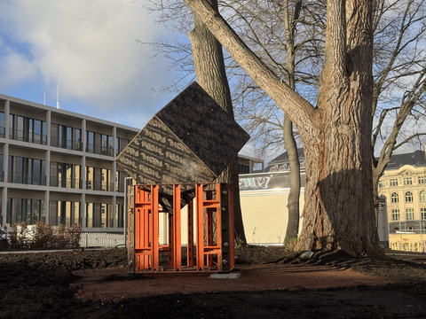 06_KSC_Collateral Sculptures_Hoernschemeyer_Equation.jpeg Sculpturale Installation aus Stahl und Holz unter Bäumen, flankiert von modernen Gebäuden.Sculptural installation made of steel and wood under trees, flanked by modern buildings.Sochařská instalace z oceli a dřeva pod stromy, lemovaná moderními budovami.Instalacja rzeźbiarska wykonana ze stali i drewna pod drzewami, otoczona nowoczesnymi budynkami.Sculpturale installatie van staal en hout onder bomen, geflankeerd door moderne gebouwen.Installazione scultorea in acciaio e legno sotto gli alberi, affiancata da edifici moderni.