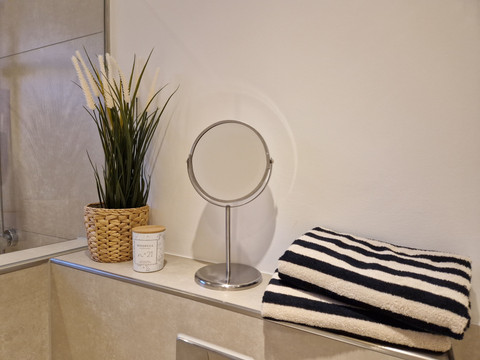 Bad_02 Runder Kosmetikspiegel auf glatter Ablage neben gestreiften Handtüchern, dekorativer Pflanze und Kerze.