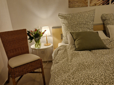 Schlafzimmer_02 Gemütliches Schlafzimmer mit beigem Bett, Blumenstrauß auf Nachttisch, Lampe und Korbstuhl.