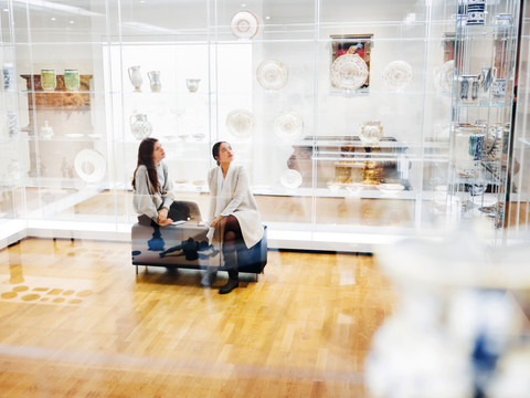 Zwei Frauen Sitzen inmitten gläserner Vitrinen mit Ausstellungsstücken wie Tellern und Kannen im GRASSI Museum für Angewandte Kunst